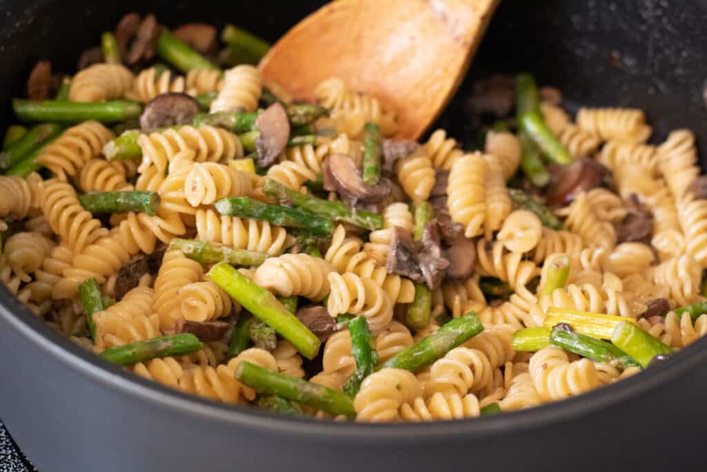 Rotini pasta with mushrooms and asparagus in a pot with a wooden spoon
