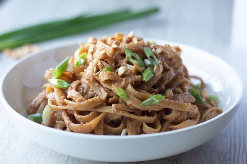 Bowl of vegetarian peanut noodles garnished with sliced green onions and chopped peanuts