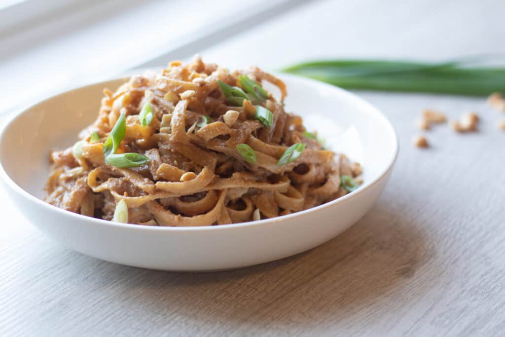 Bowl of vegetarian peanut noodles garnished with green onions and chopped peanuts