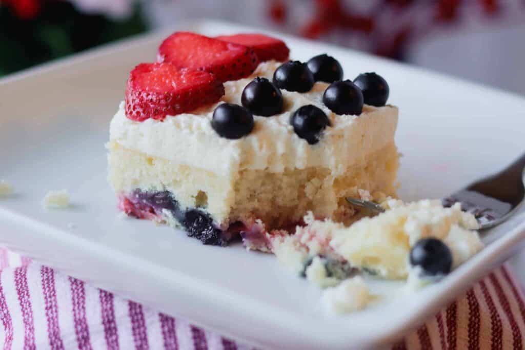 Slice of mixed berry sheet cake with whipped mascarpone frosting and topped with strawberries and blueberries.