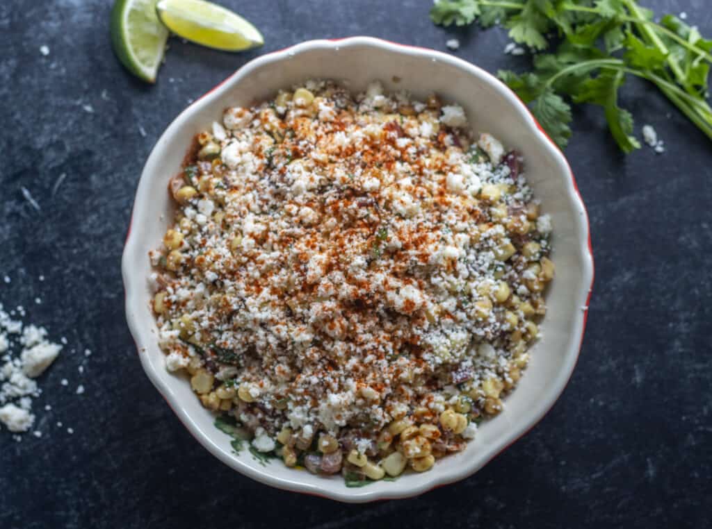 Bowl of Mexican street corn salad next to cheese, lime slices, and cilantro