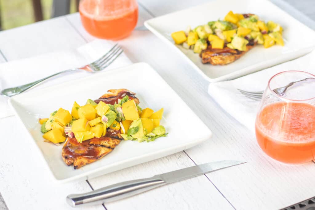 Two plates of bbq chicken topped with the mango avocado salsa on a picnic table with two glasses of strawberry lemonade