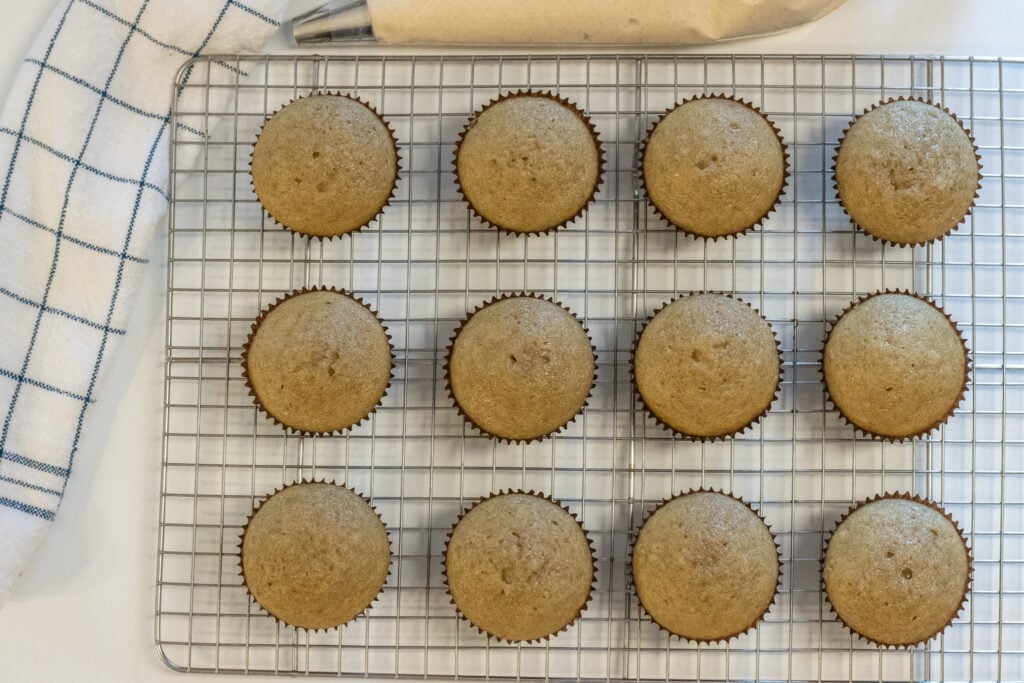 Unfrosted cardamom cupcakes on a wire rack next to a piping bag filled with frosting
