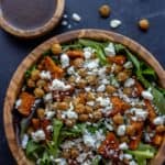 Bowl of leafy greens topped with sweet potato, chickpeas, and feta next to a bowl of balsamic dressing