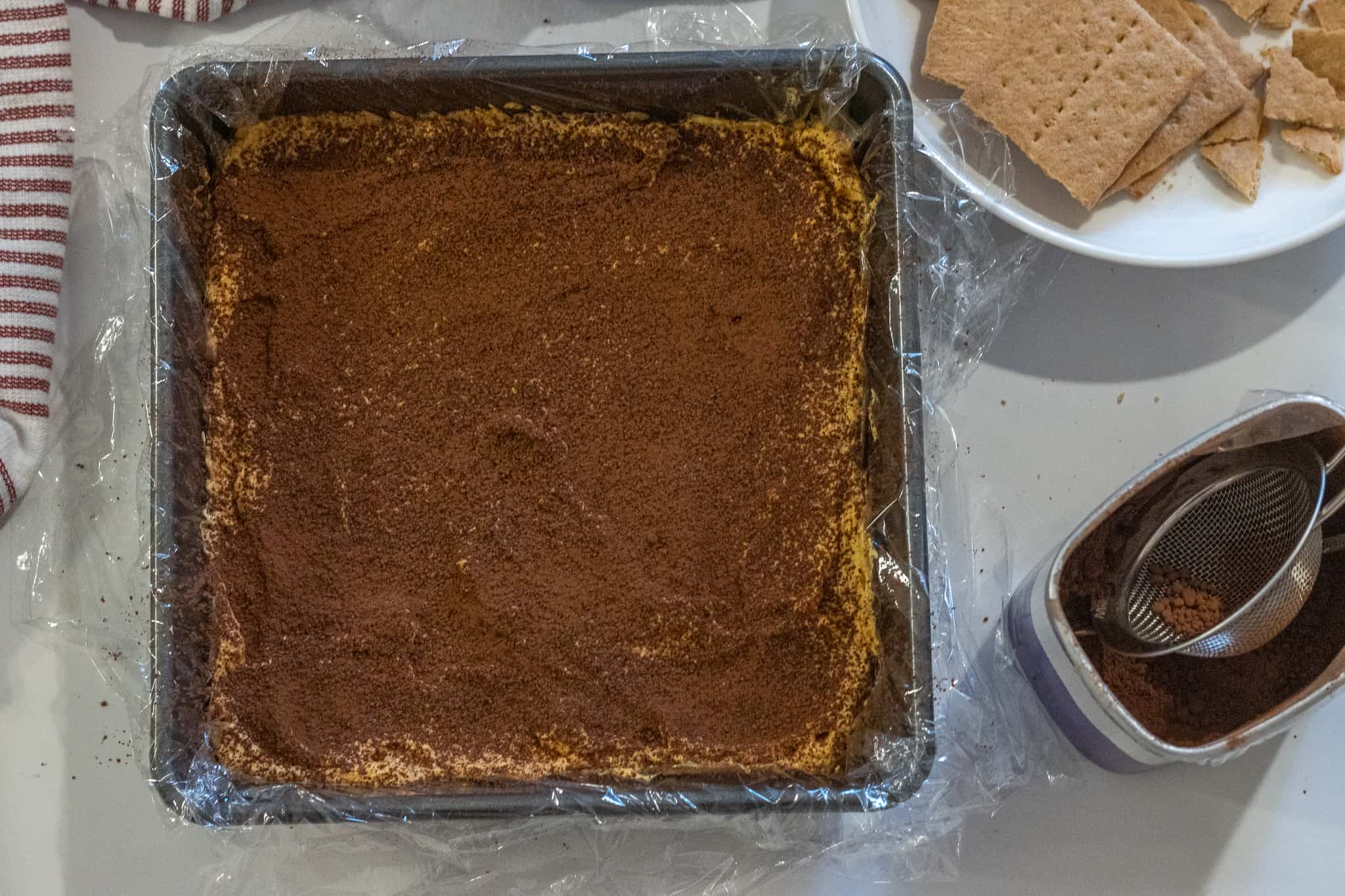 Plastic lined baking dish filled with whipped pumpkin and a layer of cocoa powder next to a plate of graham crackers and a container of cocoa powder