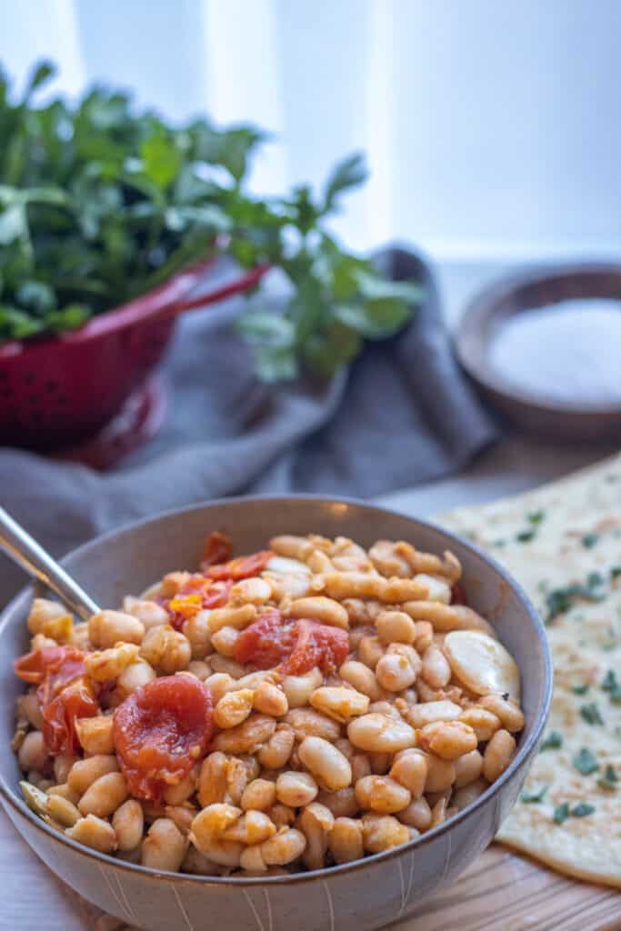 Bowl of white beans with burst tomatoes next to flatbreads with parsley and salt in the background
