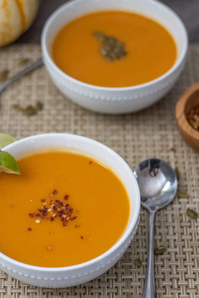 Two bowls of creamy pumpkin and coconut soup topped with red pepper flakes and pepitas