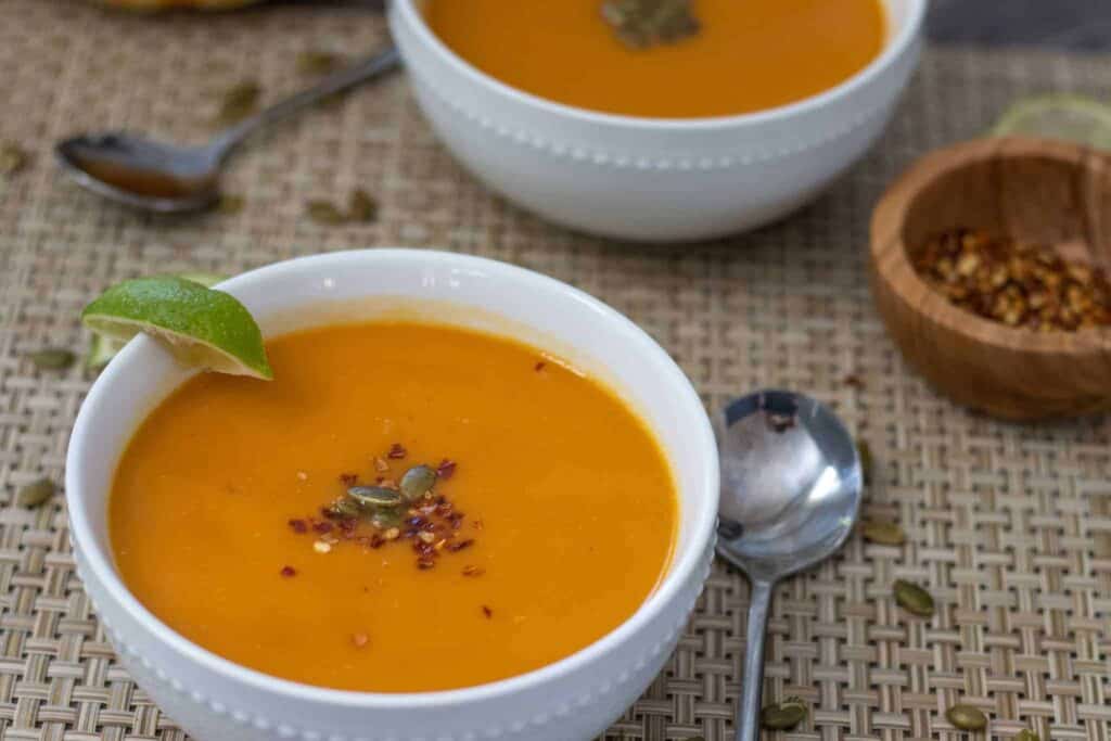 Two bowls of creamy pumpkin and coconut soup topped with red pepper flakes and pepitas