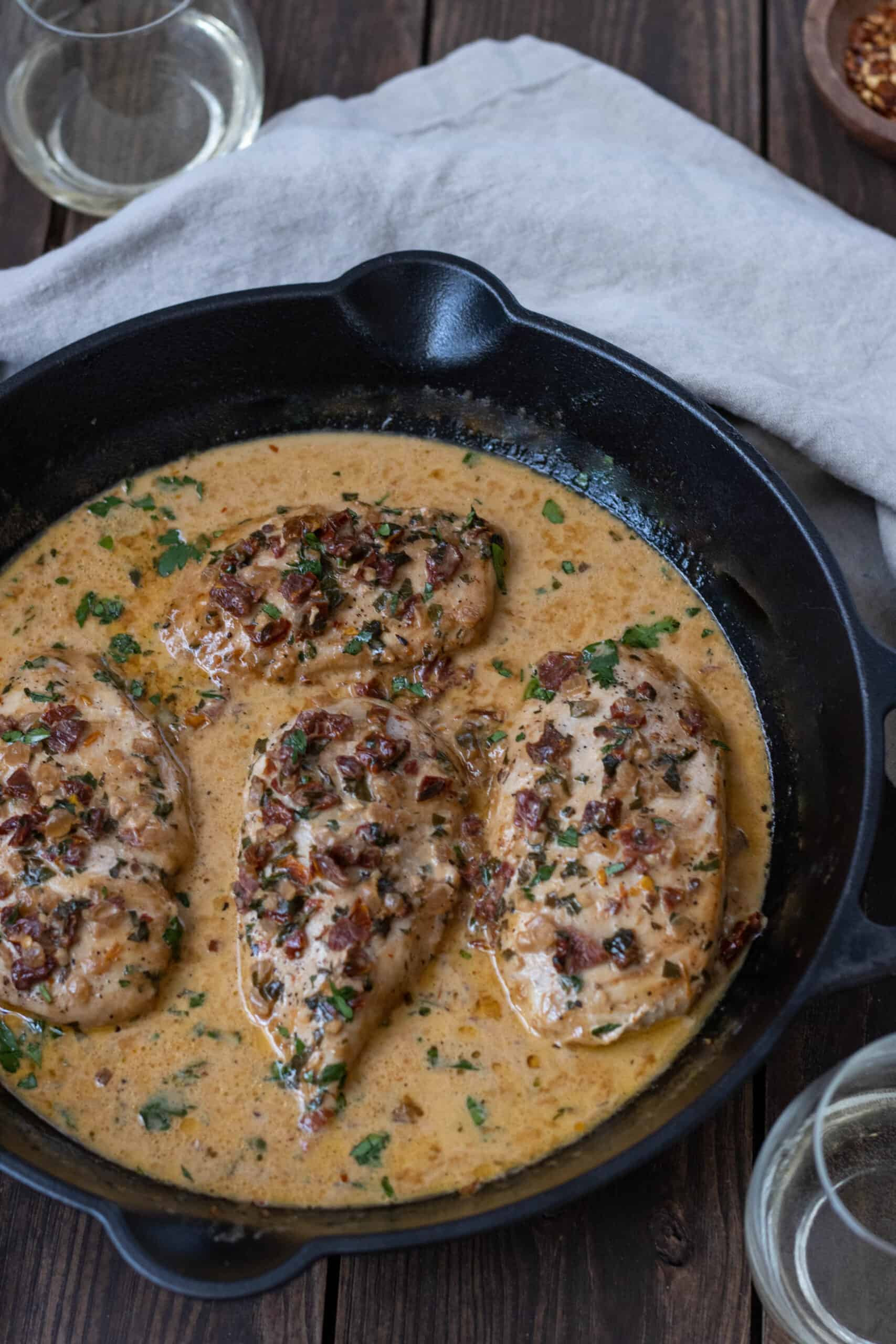 Pan with four chicken breasts in a creamy sundried tomato sauce with two glasses of white whine and a bowl with red pepper flakes