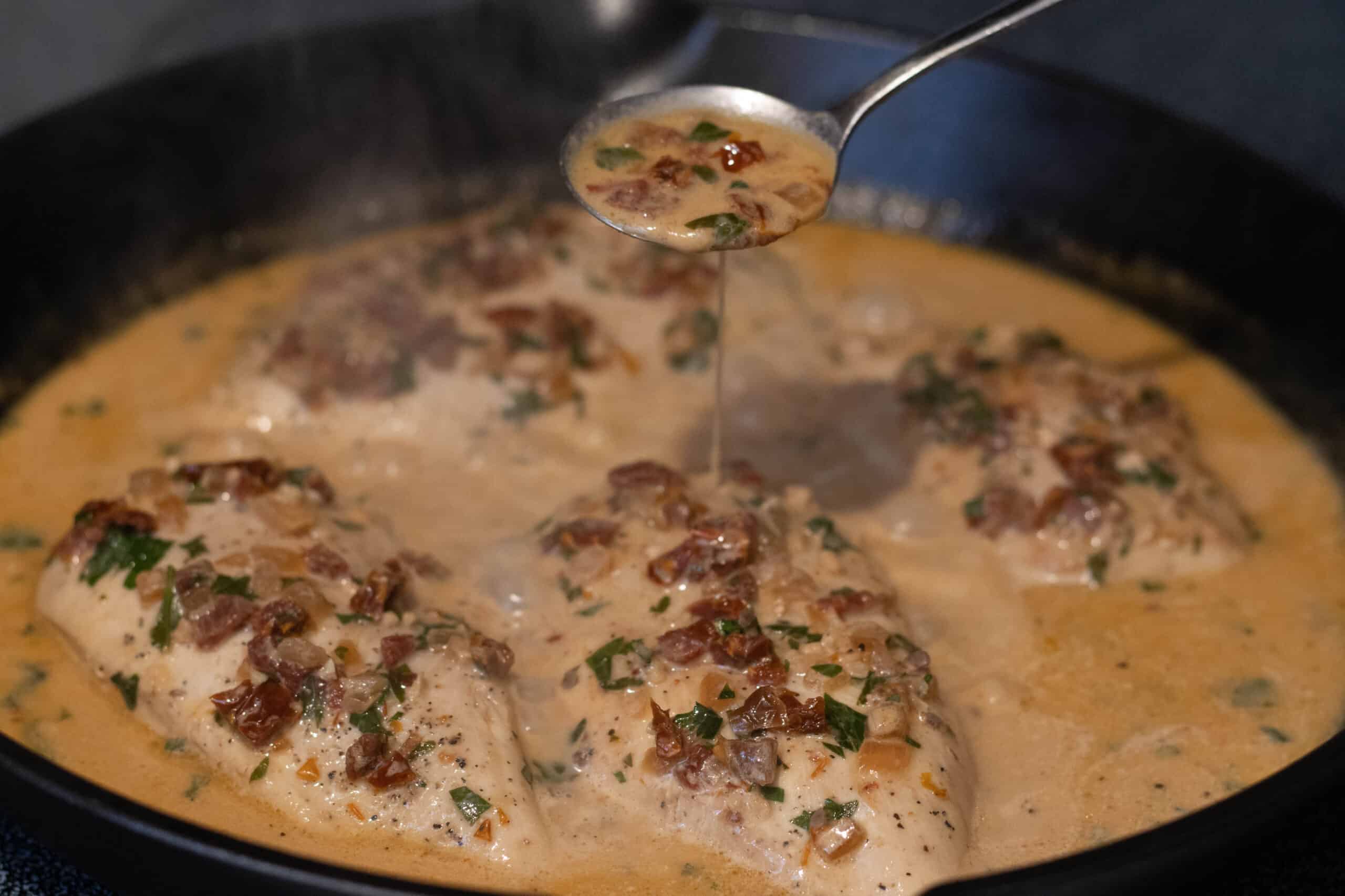 Chicken breasts cooking in creamy sundried tomato sauce in a cast iron pan with the sauce being poured over top