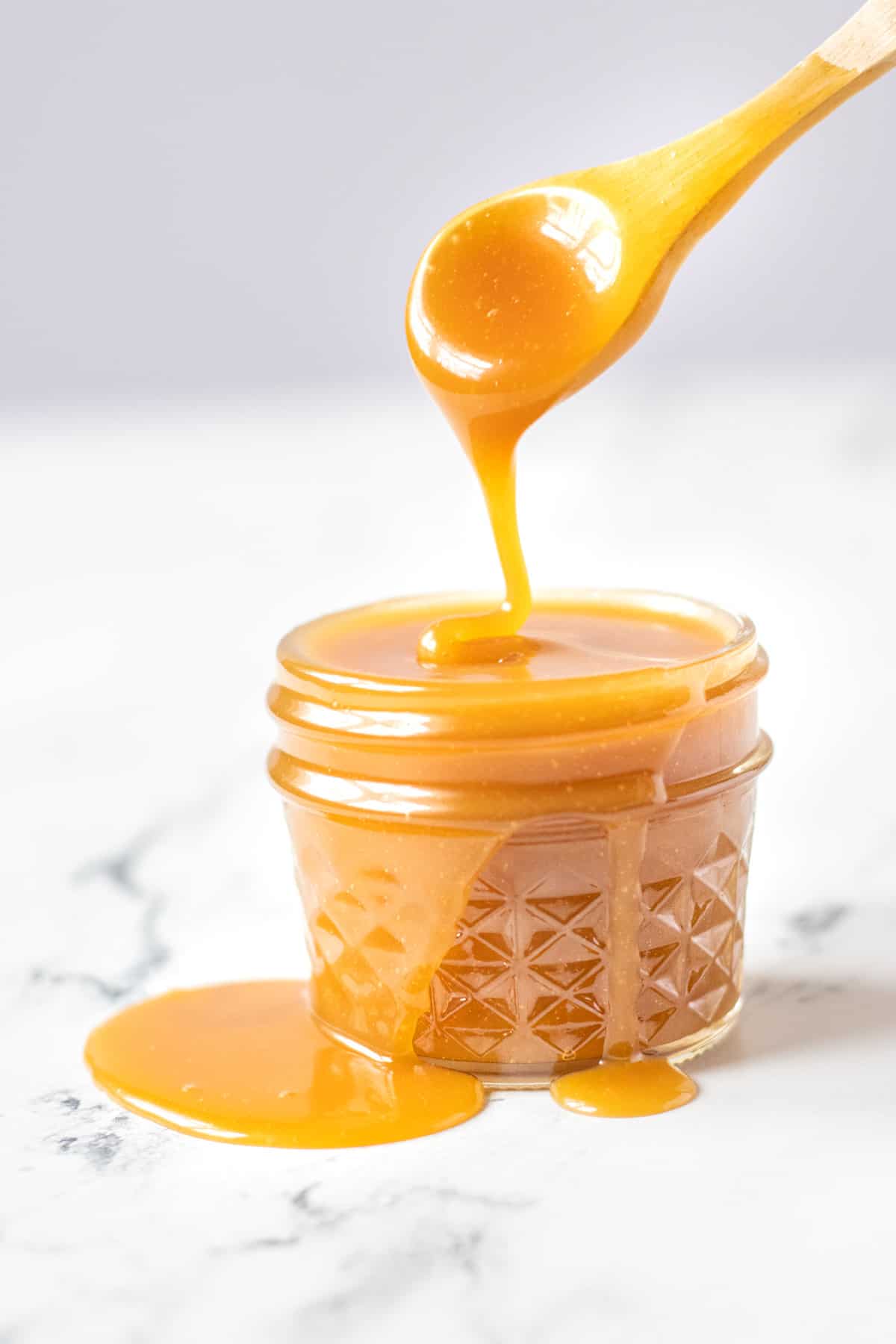 Glass jar overfilled with caramel sauce dripping over the edge with a caramel dipped wooden spoon.