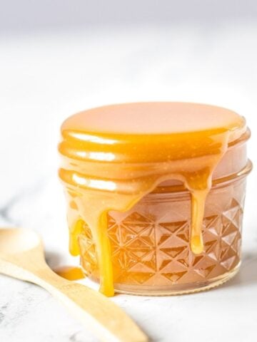 Glass jar overfilled with caramel sauce dripping over the edge with a wooden spoon next to it.