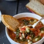 White bowl with kale and bean soup with a slice of crusty bread in it and more bread and another bowl in the background.