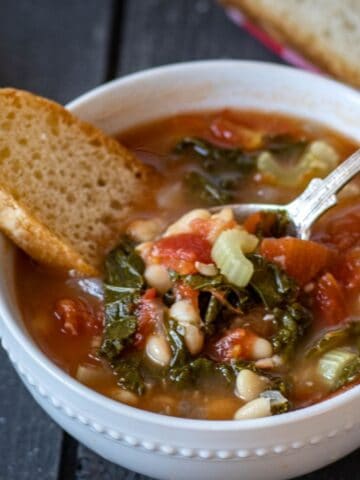 White bowl with kale and white bean soup with a slice of crusty bread and a spoon.