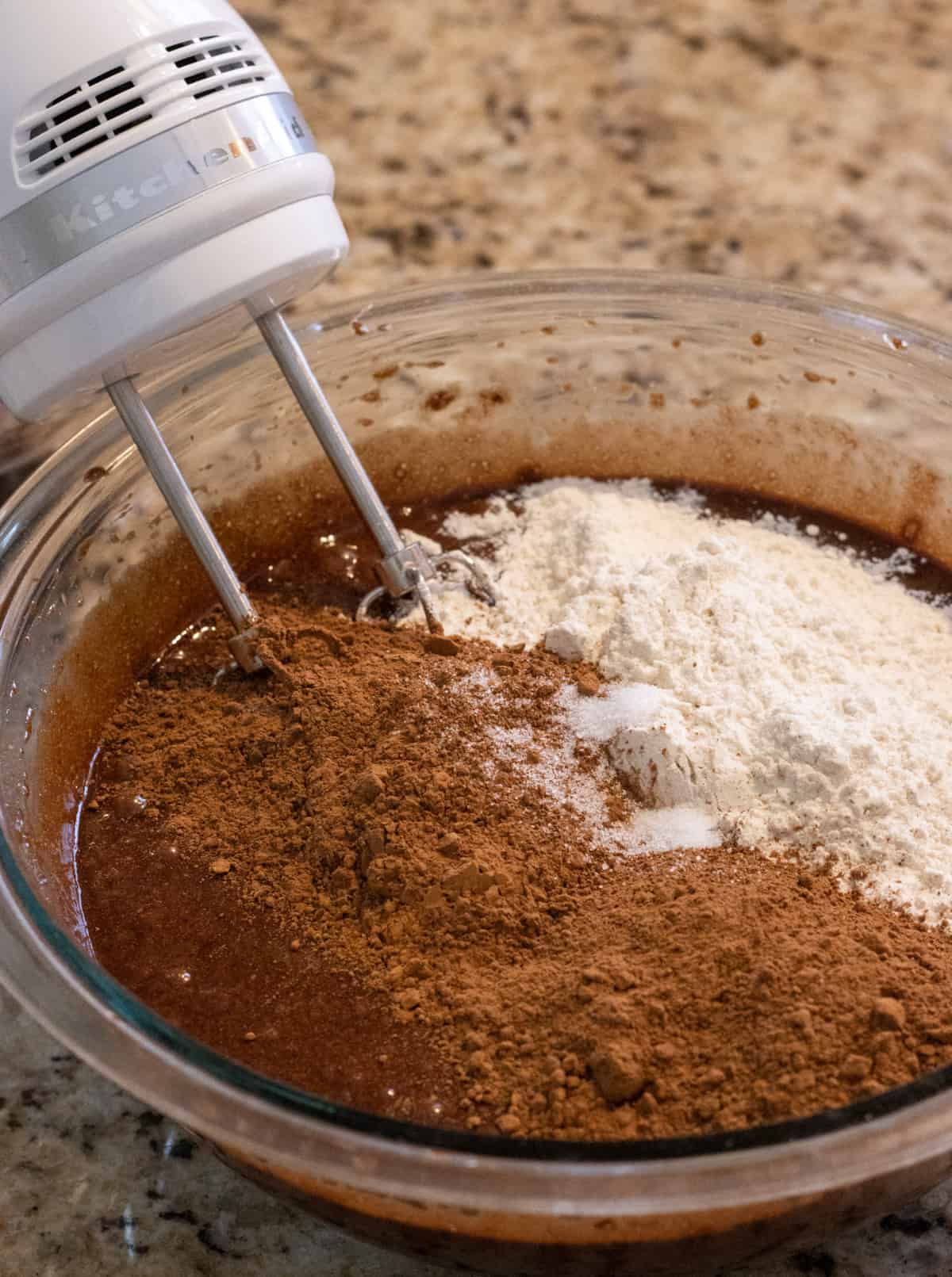 Flour and cocoa powder in a glass bowl with brownie batter and an electric whisk.