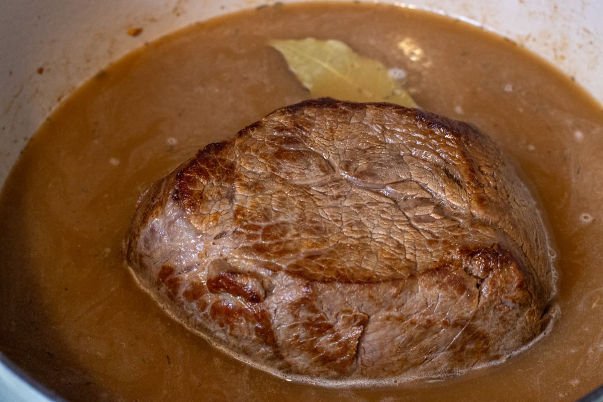 Seared chuck roast in a Dutch oven with beef broth and a bay leaf.