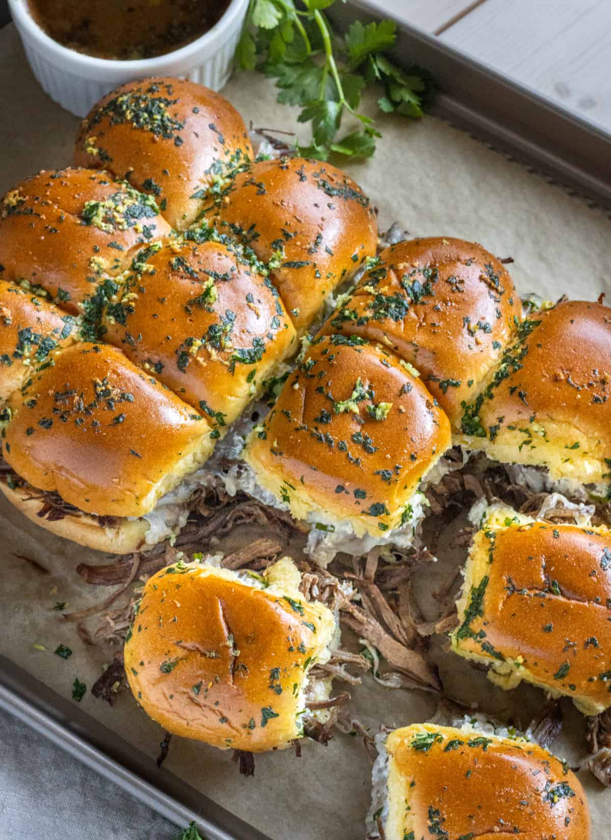 French dip sliders on a parchment lined baking sheet with a cup of au jus and parsley in the background.