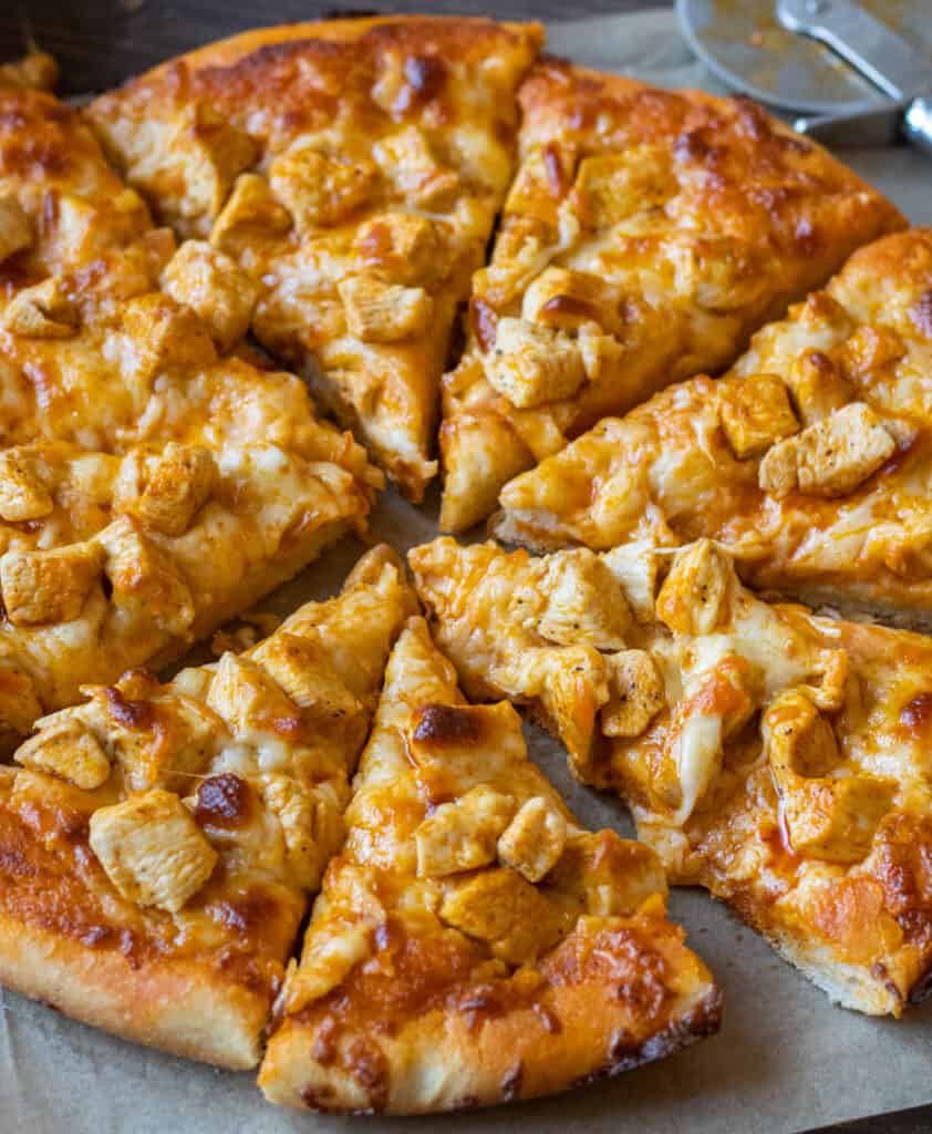 A sliced Buffalo chicken pizza on a parchment lined board with a pizza cutter in the back.