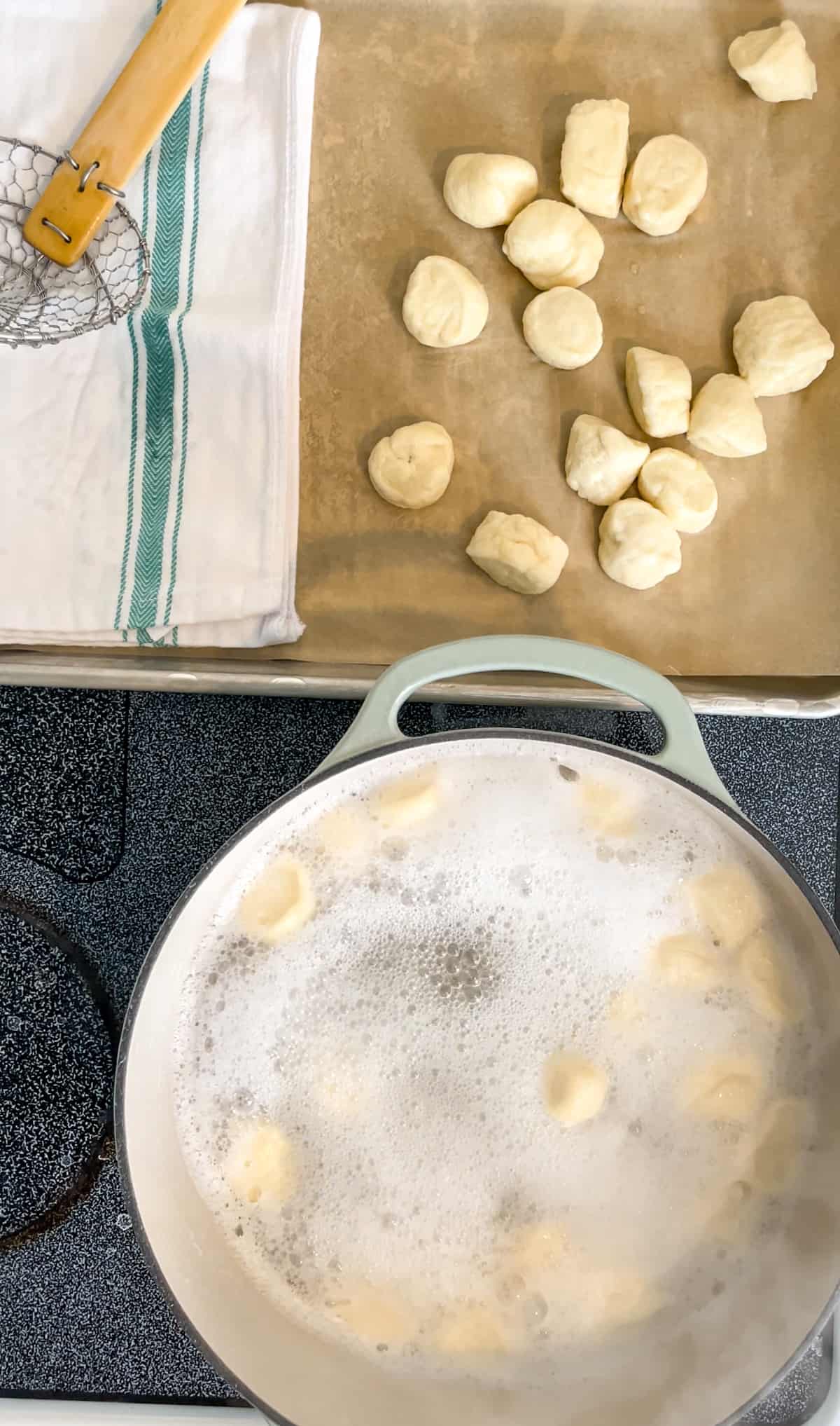 Pretzel dough pieces boiling in baking soda water and boiled pieces on a parchment-lined baking sheet with a towel and steel spider scooper.