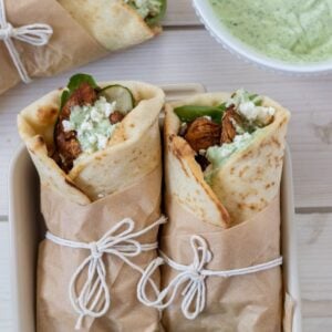 Two adobo chicken wraps wrapped in parchment and string with a third on the table and a bowl of avocado dressing.