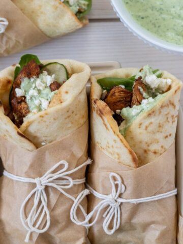 Two adobo chicken wraps wrapped in parchment and string with a third on the table and a bowl of avocado dressing.