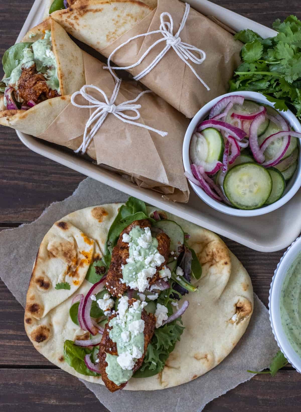 Open chicken wrap with green goddess dressing next to a tray of two wraps closed and wrapped with string next to a bowl of cucumbers and red onions.