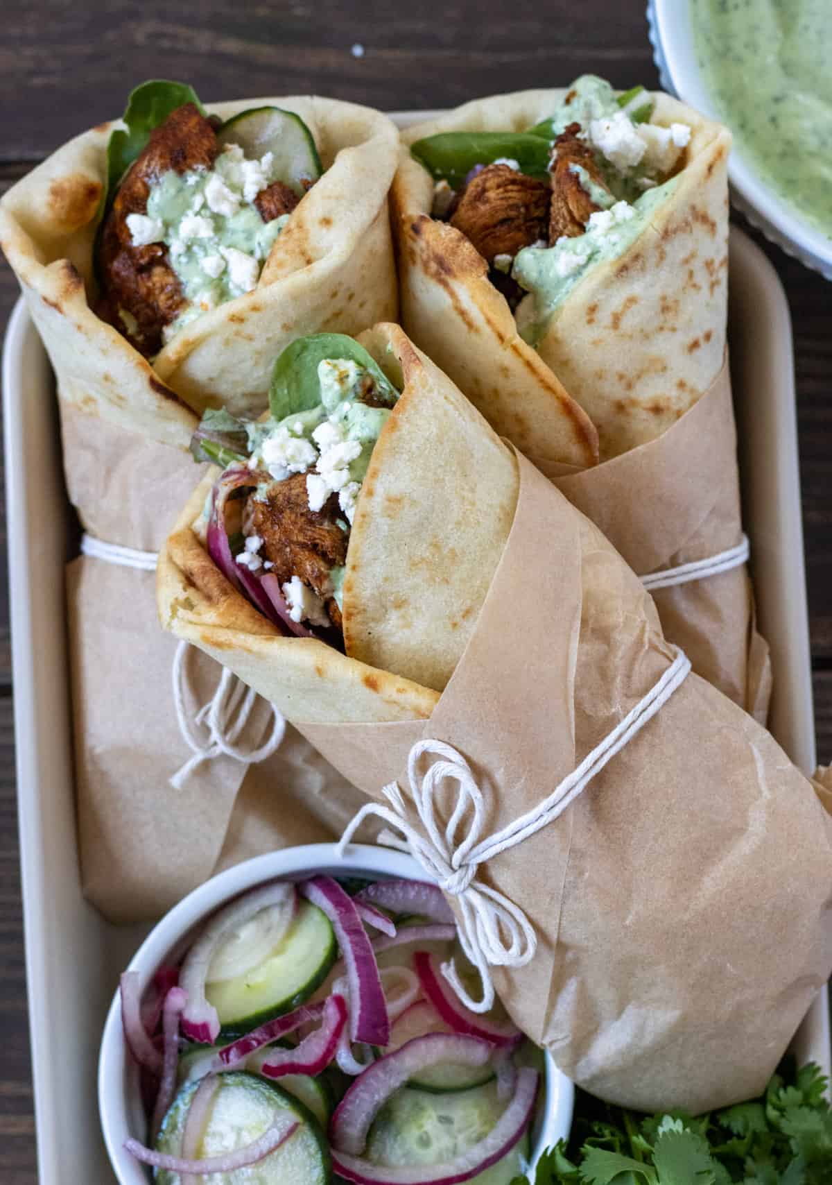 Three chicken and naan wraps wrapped in parchment and string with a bowl of sliced cucumbers and red onion.