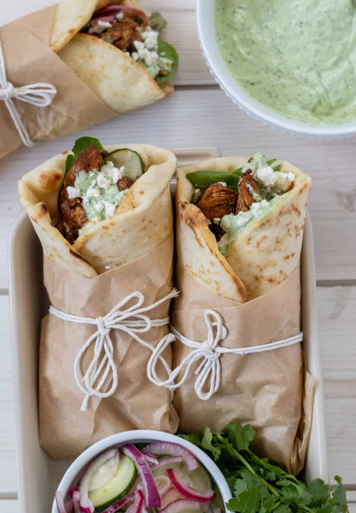 Two chicken wraps in a tray with a bowl of sliced cucumbers and red onions with a third wrap on the table and a bowl of avocado dressing on the side.