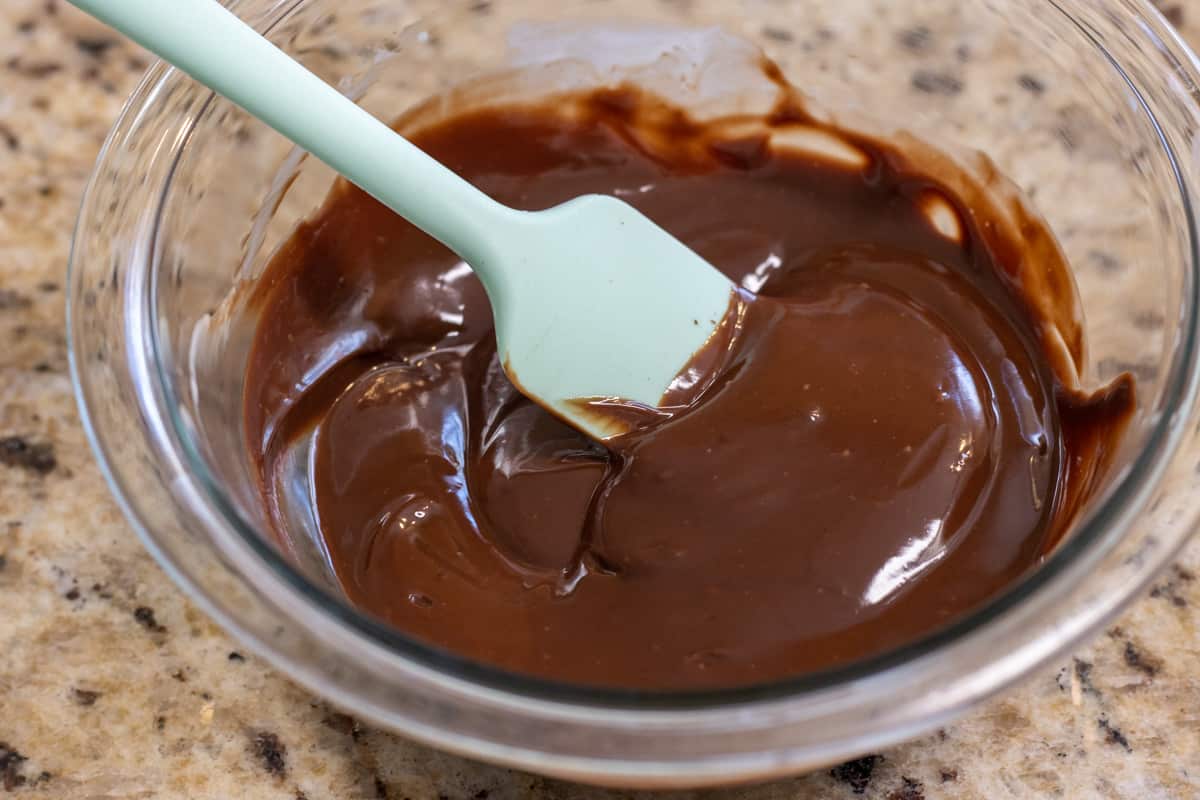 Chocolate ganache in a glass bowl with a blue rubber spatula.