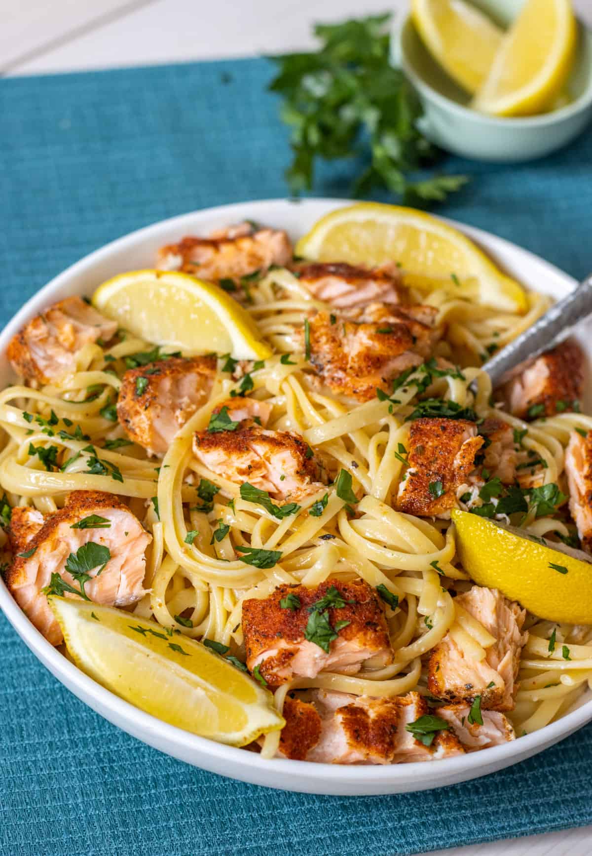 Linguine topped with chunks of salmon, lemon wedges, and parsley with extra parsley and lemons in the background.