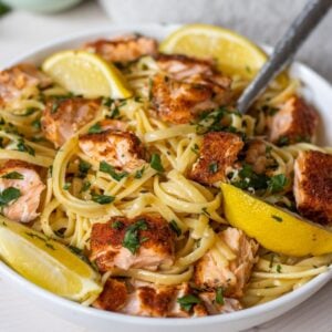 Bowl of linguine pasta with chunks of seasoned salmon and lemon wedges with a fork sticking out.