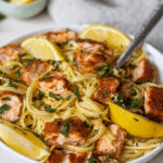 Bowl of linguine with chunks of seasoned salmon and lemon wedges with a napkin and lemon wedges in the background.