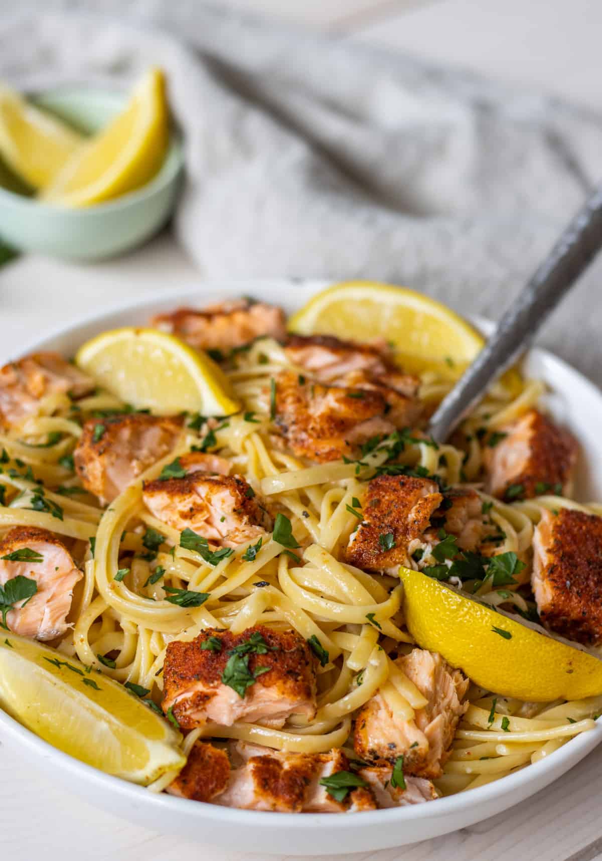 Linguine pasta in a bowl with chunks of seasoned salmon and lemon wedges with a cloth napkin and a bowl of lemon wedges in the background.