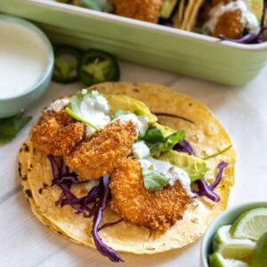 Two corn tortillas stacked up topped with three fried shrimp, purple cabbage, avocado slices, and crema with more tacos in the background.