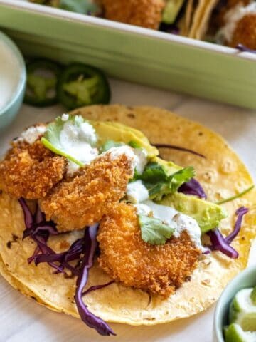 Two corn tortillas stacked up topped with three fried shrimp, purple cabbage, avocado slices, and crema with more tacos in the background.
