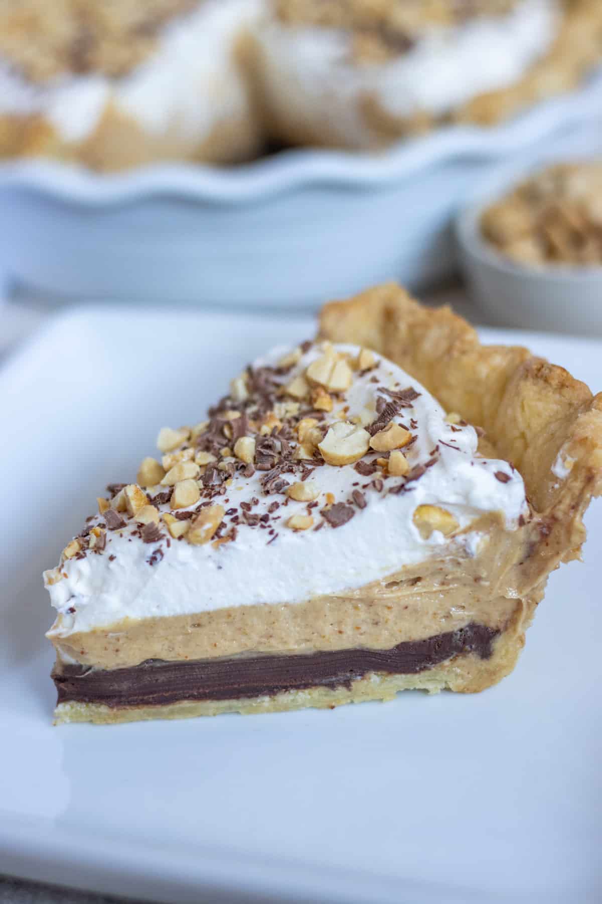 Side shot of a slice of pie with a chocolate layer, a peanut butter layer, and a whipped cream layer with chocolate shavings and chopped peanuts on top.