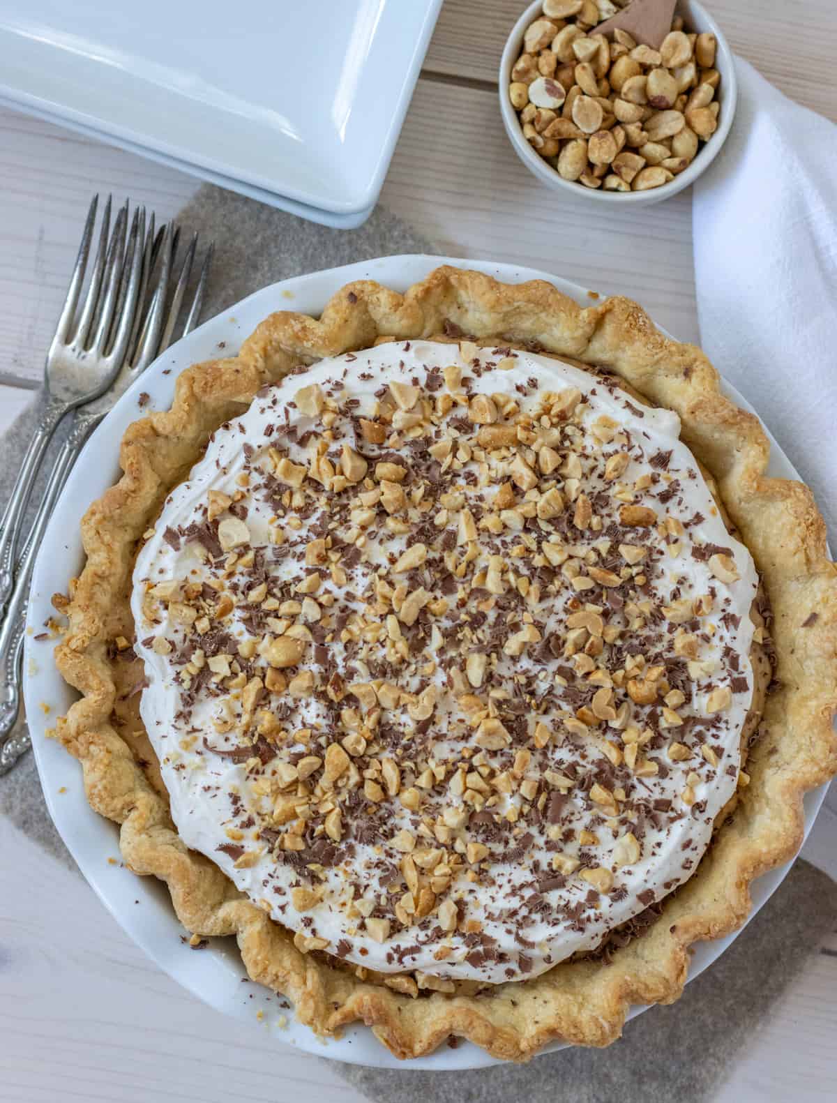 Overhead view of a peanut butter pie topped with whipped cream, chocolate shavings, and peanuts with forks, a plate and bowl of peanuts off to the side.