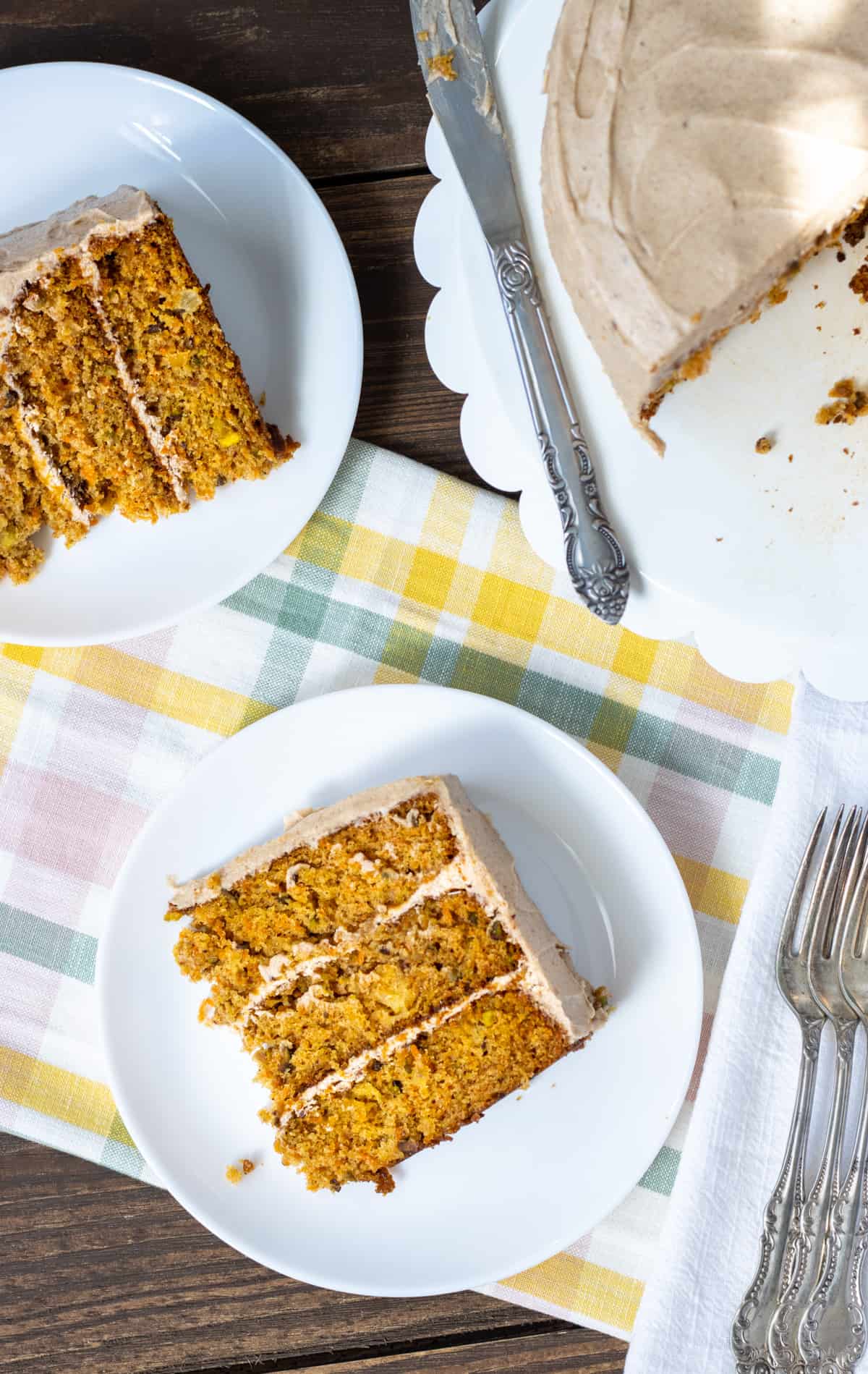 Carrot cake slices on two plates with the whole cake in the background with forks to the side.