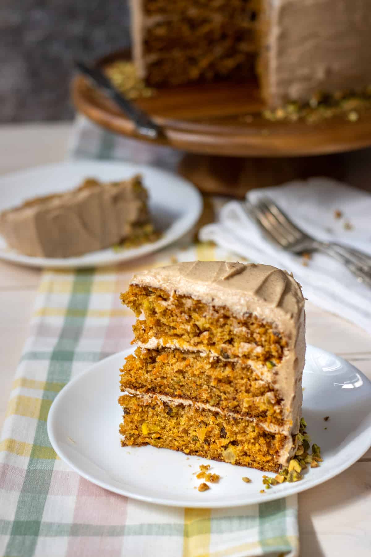 A slice of pistachio carrot cake standing up in front with another slice in the back with the full cake of a stand.