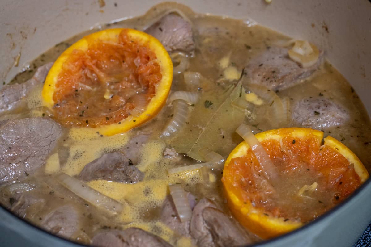 Dutch oven with pork in cooking liquid with a bay leaf and orange halves simmering.
