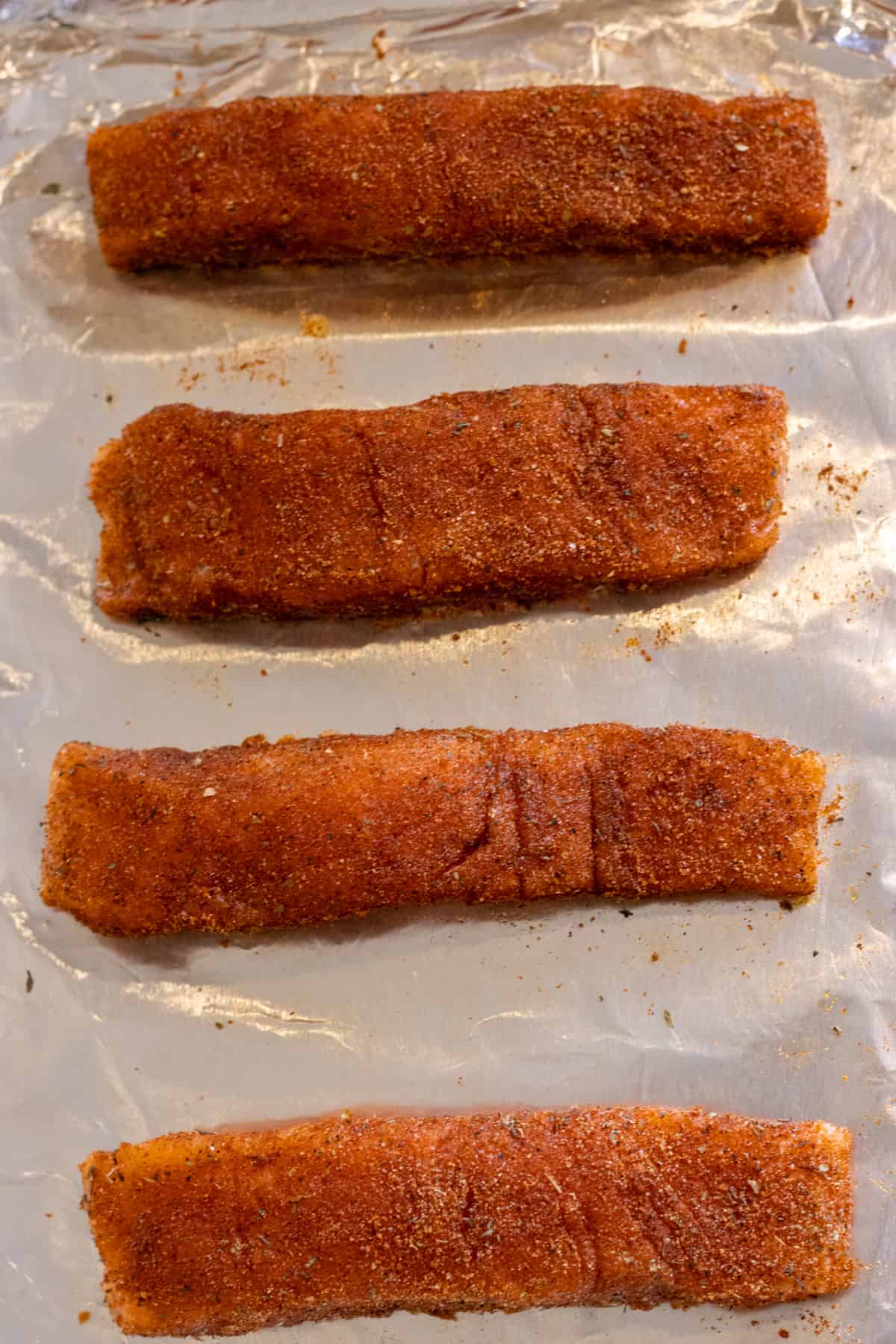 Four uncooked salmon filets coated in a blackening seasoning on a foil-lined baking sheet.