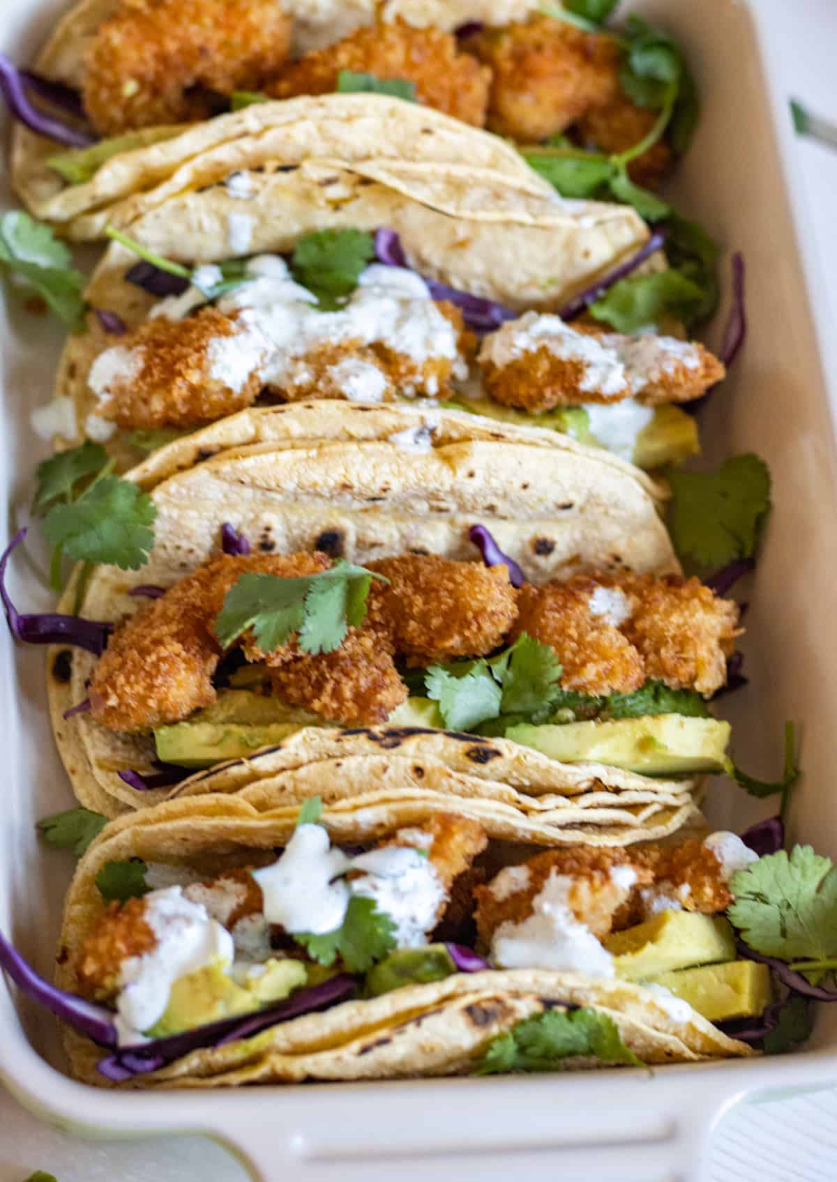 A row of fried shrimp tacos with avocado slices, purple cabbage, and crema.