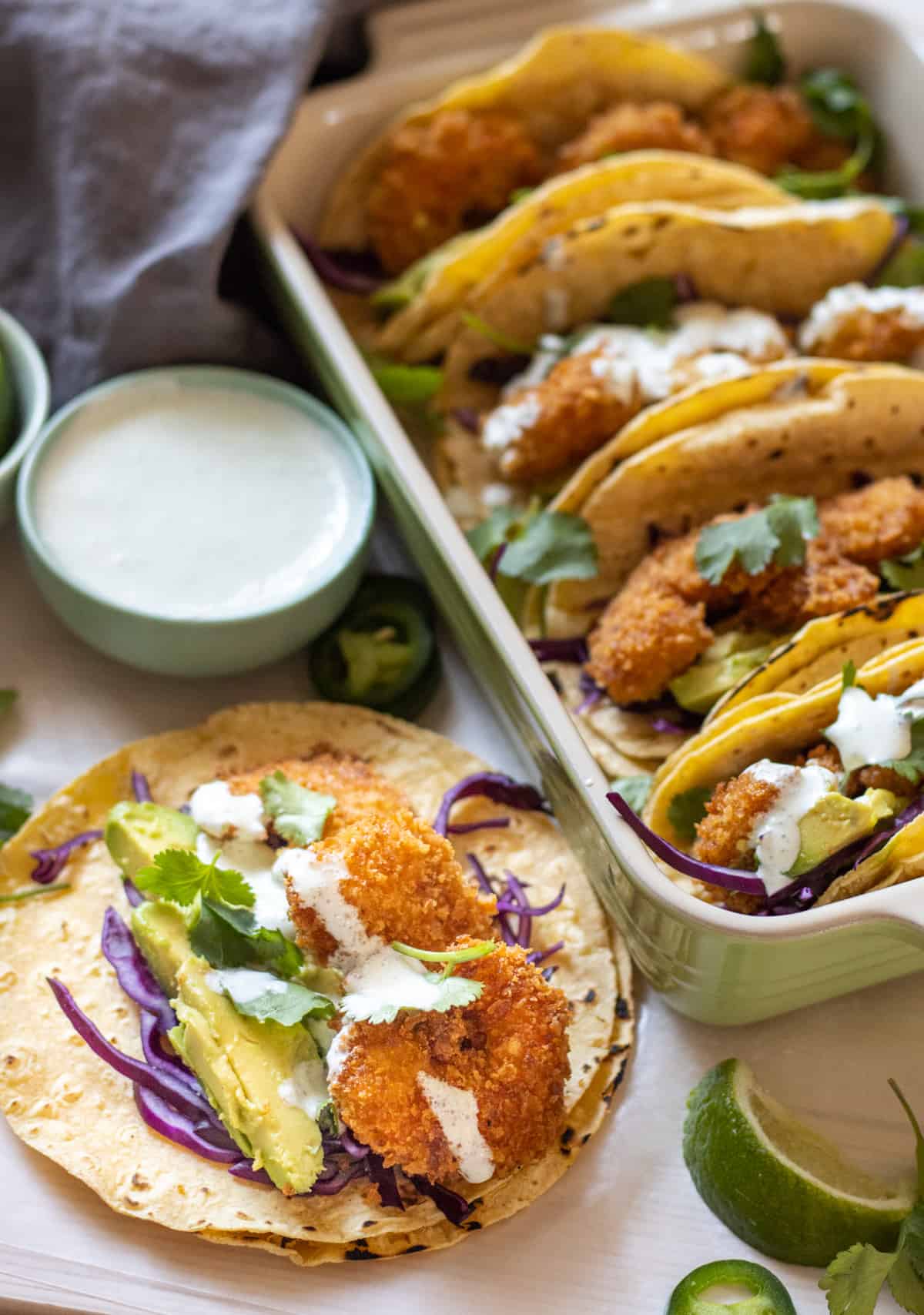 Fried shrimp tacos with purple cabbage and avocado slices lined up in a baking dish with another one laid open next to them.