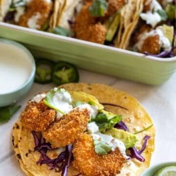 Fried shrimp taco with red cabbage, avocado, and crema on board with a tray of tacos in the background.