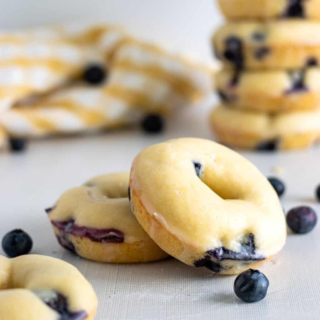 Blueberry donuts on a table with blueberries and more donuts stacked in the background.