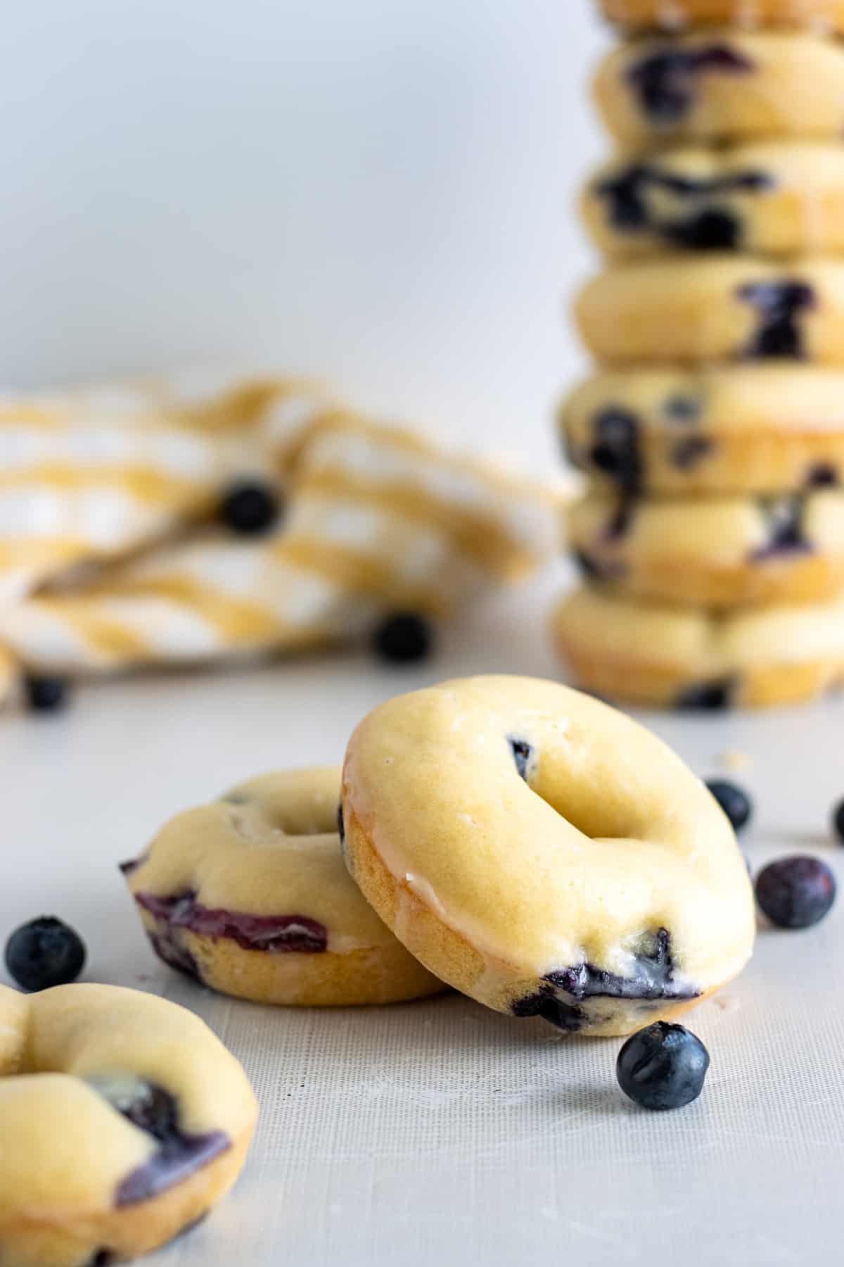 Glazed blueberry donuts and blueberries on a table with a stack of donuts and a yellow towel in the background.