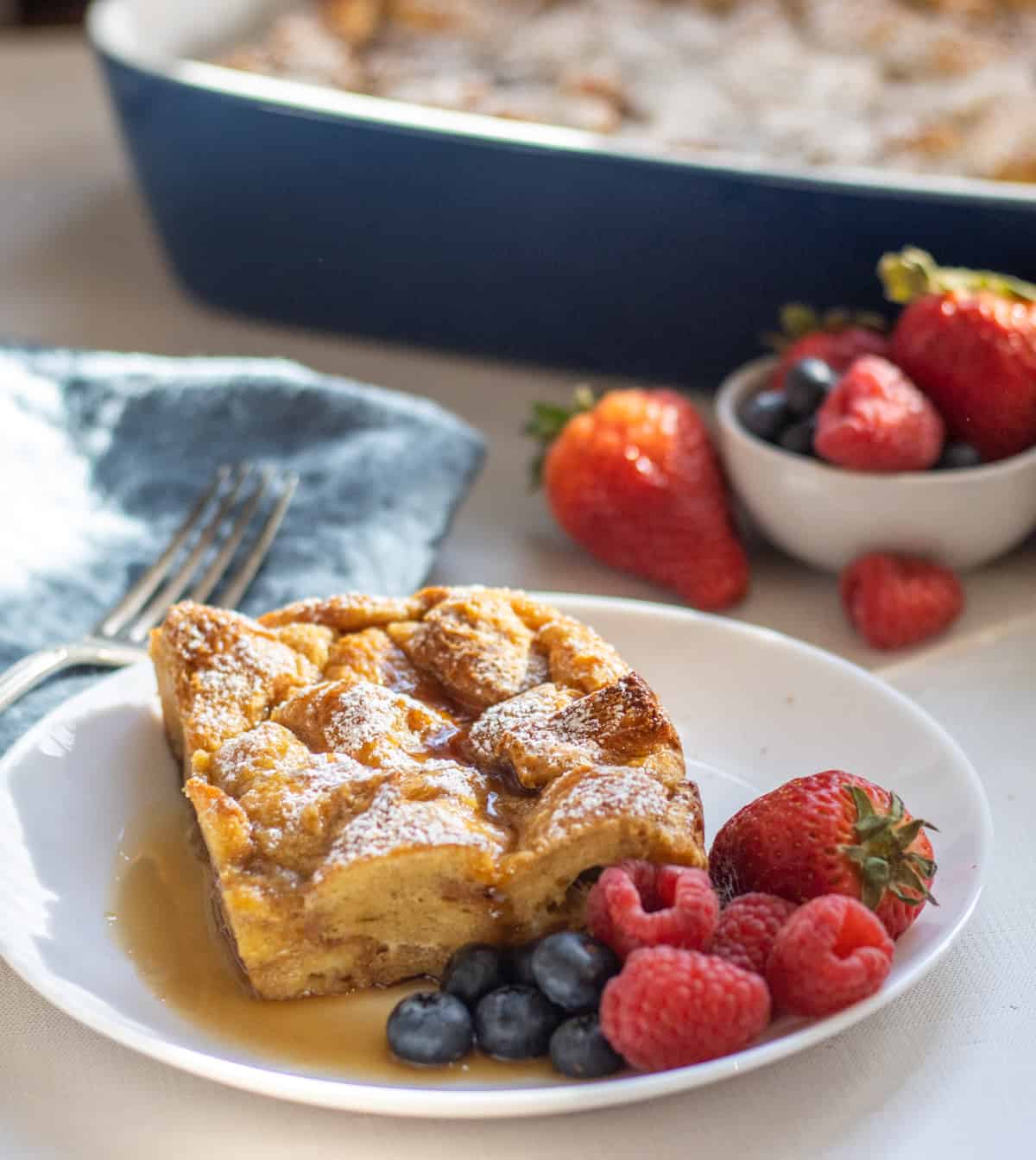 Slice of French toast casserole on a plate with mixed berries and the baking dish in the background next to more berries.