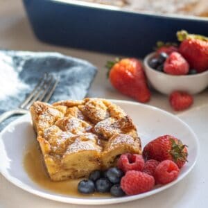 Slice of French toast casserole on a plate with mixed berries and maple syrup with more berries and the casserole in the background.