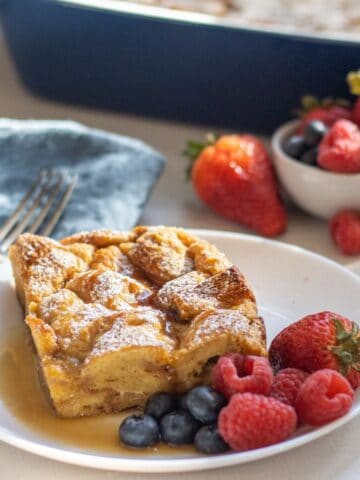 Slice of French toast casserole on a plate with mixed berries and maple syrup with more berries and the casserole in the background.