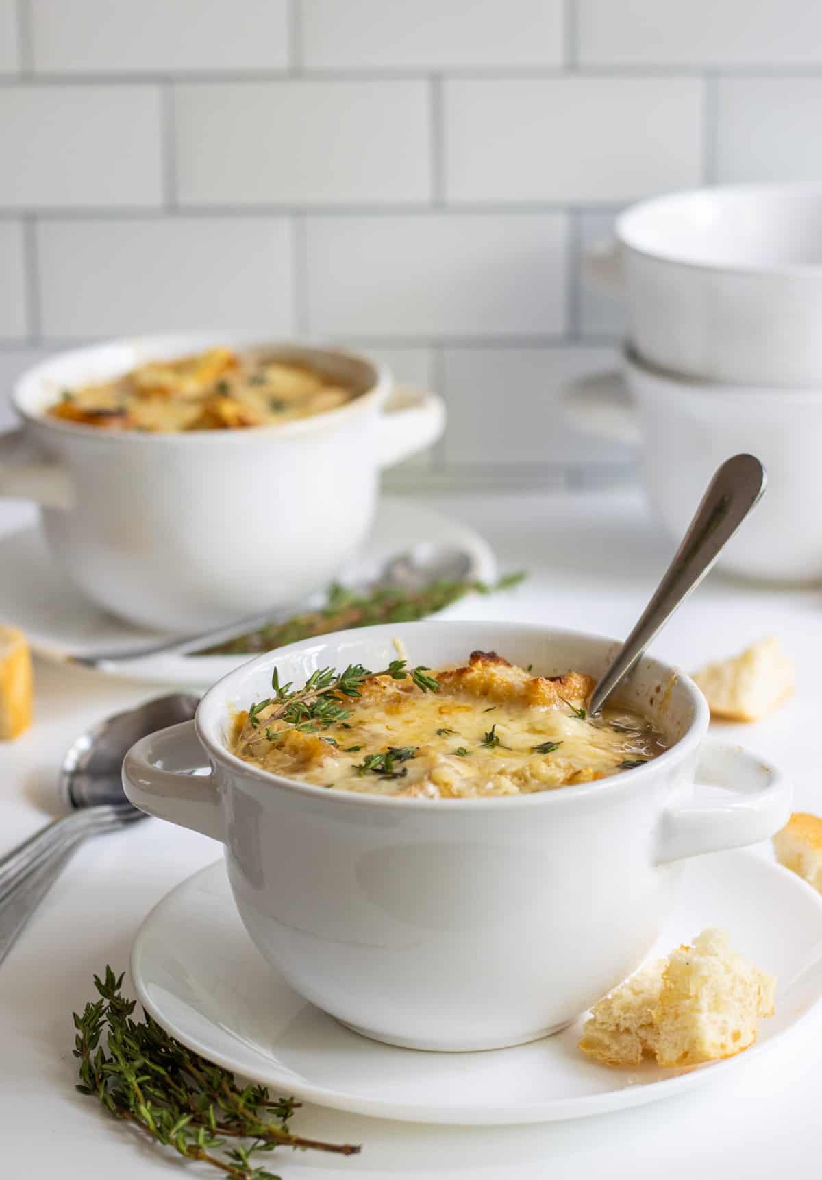 Two bowls of French onion soup with sprigs of thyme and bread cubes all around with extra bowls in the background.